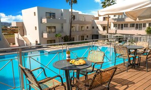 Ett område vid poolen med flätade stolar och bord, en klarblå pool och moderna, vita byggnader i bakgrunden.