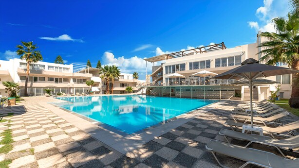 A swimming pool with lounge chairs and umbrellas, surrounded by palm trees and a building with multiple balconies. - The Garden Studio Pool View (Chania)