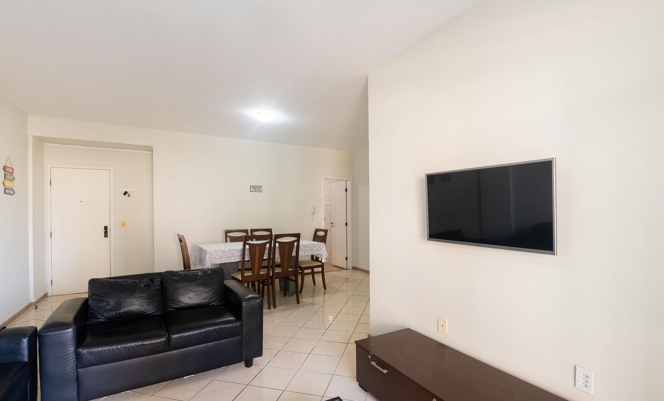 A living room with a black leather sofa, a dining table with chairs, and a flat-screen TV mounted on the wall.