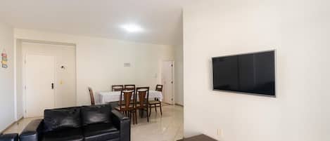 A living room with a black leather sofa, a dining table with chairs, and a flat-screen TV mounted on the wall.