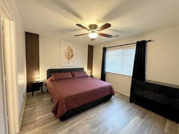 Une chambre à coucher comprenant un lit, un ventilateur de plafond, une fenêtre avec des stores et un téléviseur fixé au mur.