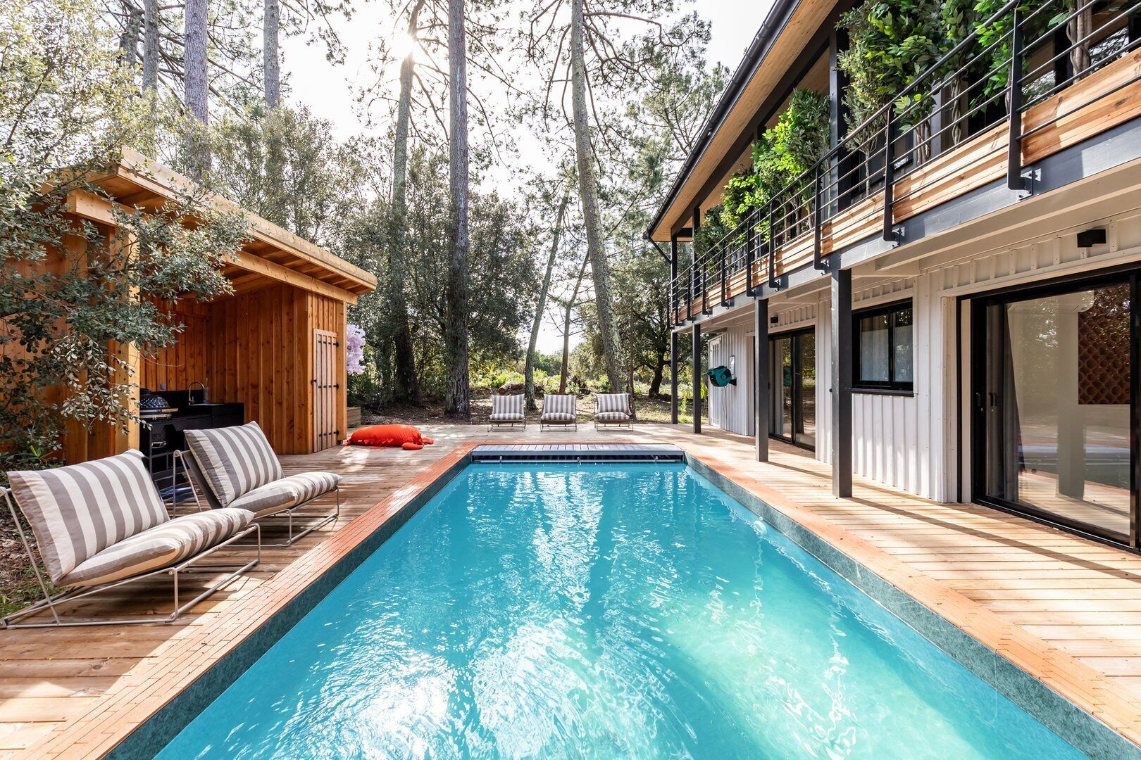Un espace piscine extérieur moderne, doté d’une terrasse en bois, d’un barbecue intégré et d’un coin salon avec des coussins à rayures.