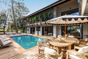 A modern outdoor pool area with a wooden deck, lounge chairs, and a dining table with umbrellas. - Villa Domus Sciurorum – Pyla-sur-Mer (Pyla sur Mer)