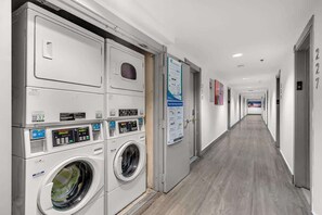 A hotel laundry room with multiple washing machines and dryers, a long corridor with doors numbered 227, and informational posters on the walls. - Oceanfront Studio · South of Fifth + Beach Perks (Miami Beach)