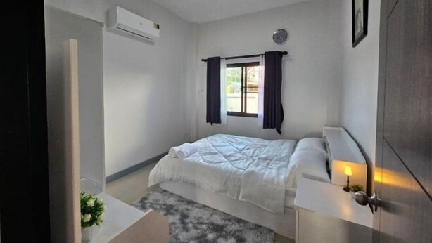 Een slaapkamer met een bed, een raam met gordijnen, een airconditioningapparaat, een nachtkastje met een lamp en een klok aan de muur.