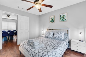 Une chambre à coucher comprenant un lit, un ventilateur de plafond, une table de chevet, un bureau avec des chaises et deux tableaux encadrés accrochés au mur.