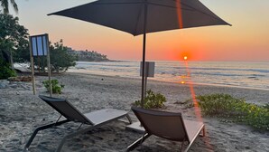 Two sun loungers with a beach umbrella, a chair, and a small table on a sandy beach with a sunset view. - Luxury Oceanfront At Majahua (Troncones)