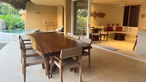 A spacious dining area with a large wooden table and chairs, a glass-enclosed patio with a view of greenery, and a cozy seating area with a sofa and chairs. - Luxury Oceanfront At Majahua (Troncones)