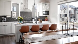 Una cocina moderna con armarios blancos, una isla central con taburetes y un amplio ventanal con vista a la montaña, además de iluminación colgante.
