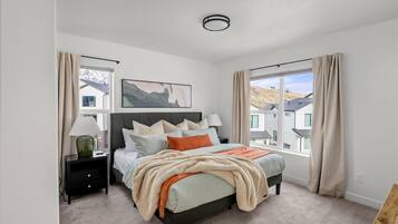 Un dormitorio con cama, mesitas de noche, lámpara, un cuadro y vistas a casas.