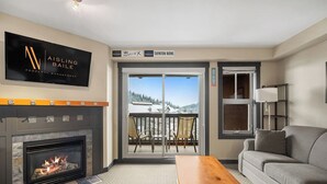 Ein Wohnzimmer mit Kamin, einer Couch, einem Couchtisch und einem an der Wand befestigten Fernseher. Eine Glastür öffnet sich zu einem Balkon mit Blick auf eine verschneite Landschaft.