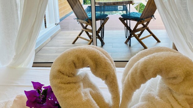 Een kamer met uitzicht op een houten terras en groen, met een handdoekje gevouwen in de vorm van een hart en een tafel met stoelen.