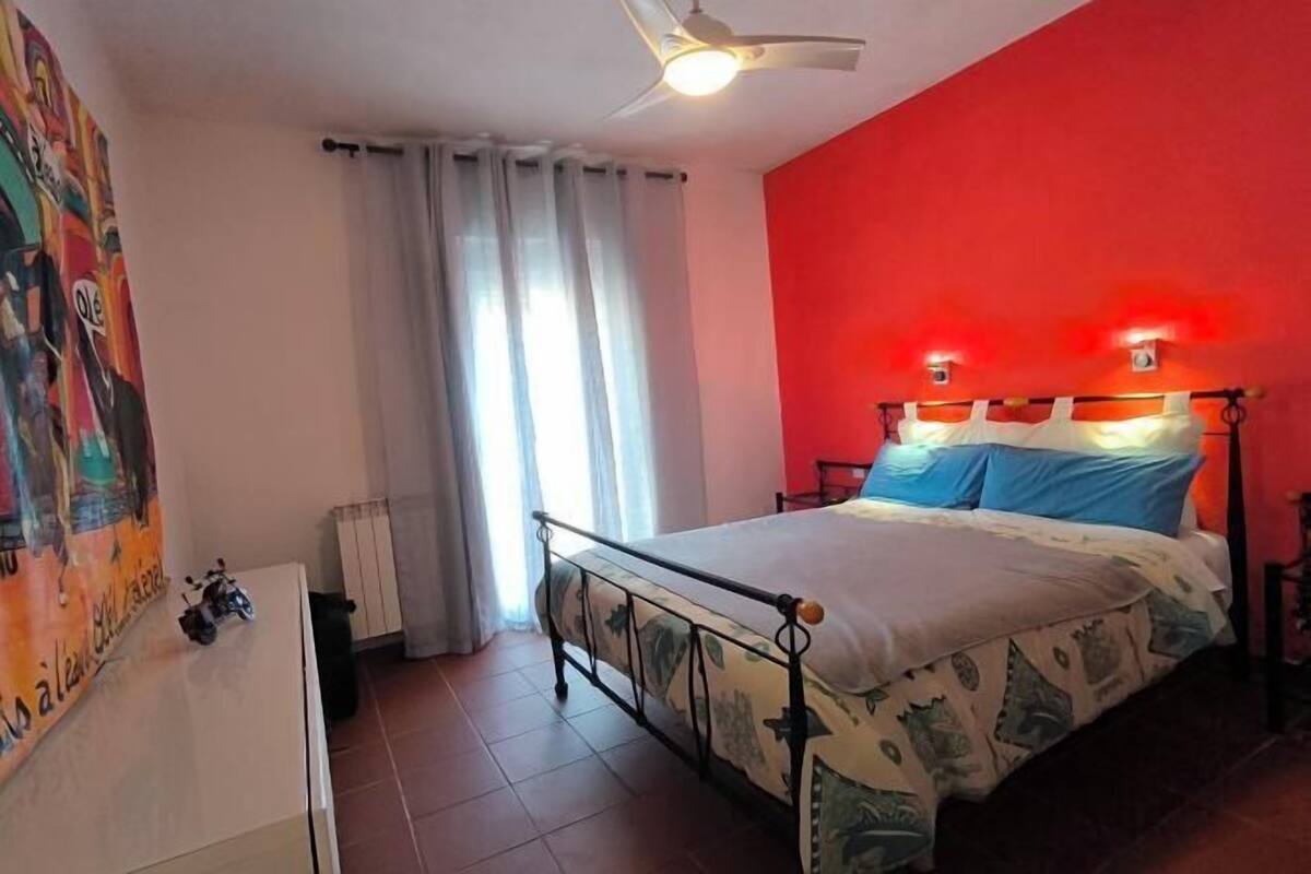 Une chambre à coucher comprenant un lit, un ventilateur de plafond, un radiateur, une fenêtre avec des rideaux et un mur avec un détail décoratif rouge.