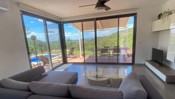 A modern living room with a large sofa, a coffee table, and a TV. There is a sliding glass door leading to an outdoor patio with a pool and chairs.