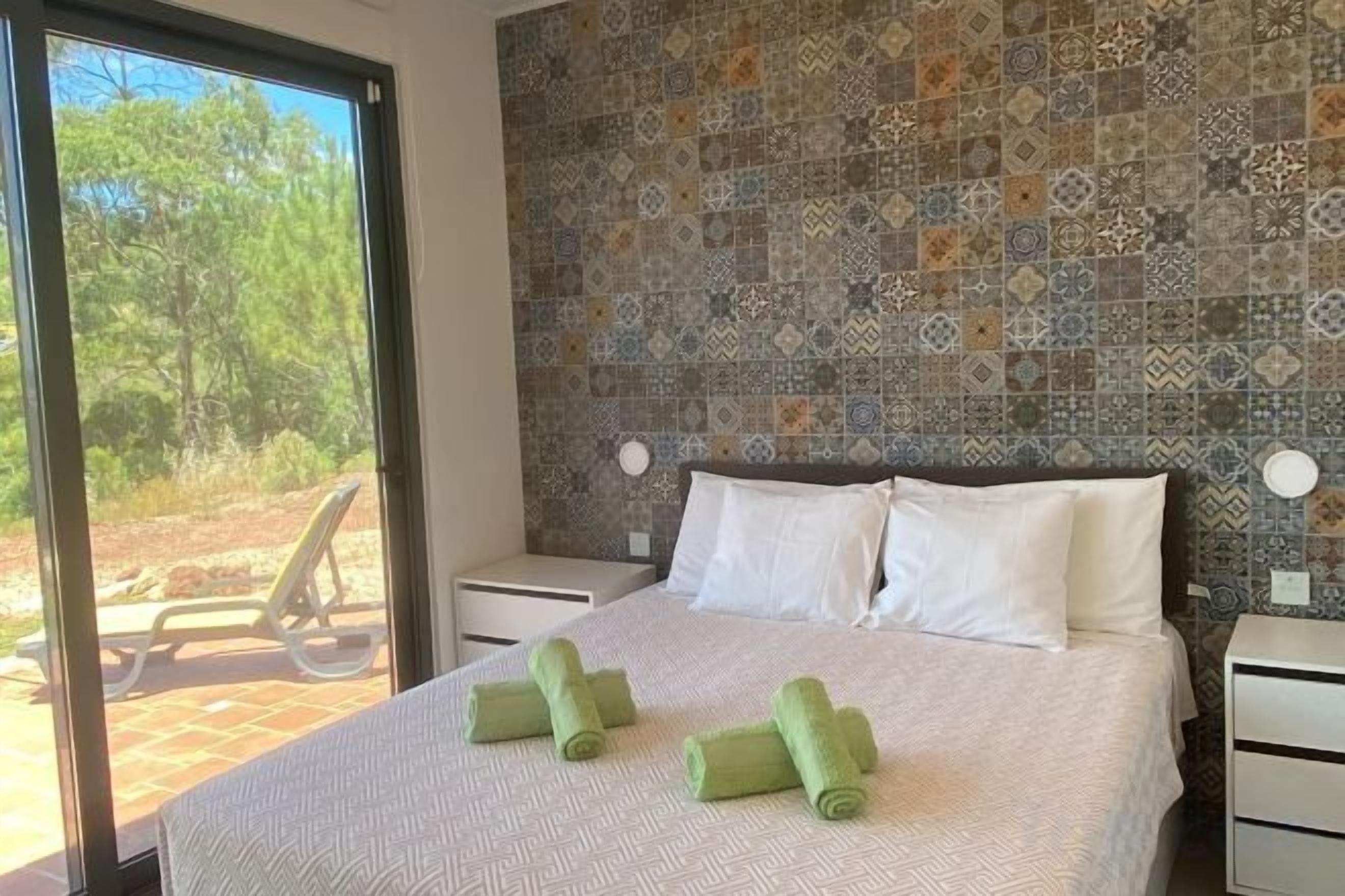 A bedroom with a patterned wall, a bed with white linens, and a view of the outdoors.