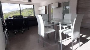 A modern office space with a glass table and white chairs, large windows with a view of greenery, and a ceiling fan.