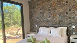 A hotel room with a patterned tile wall, a bed with white linens, and a view of greenery through the window.