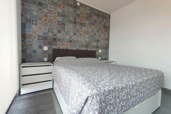 Une chambre avec un mur recouvert de carreaux à motifs, un lit avec une couvre-lit grise à motifs et une table de chevet blanche avec des tiroirs.