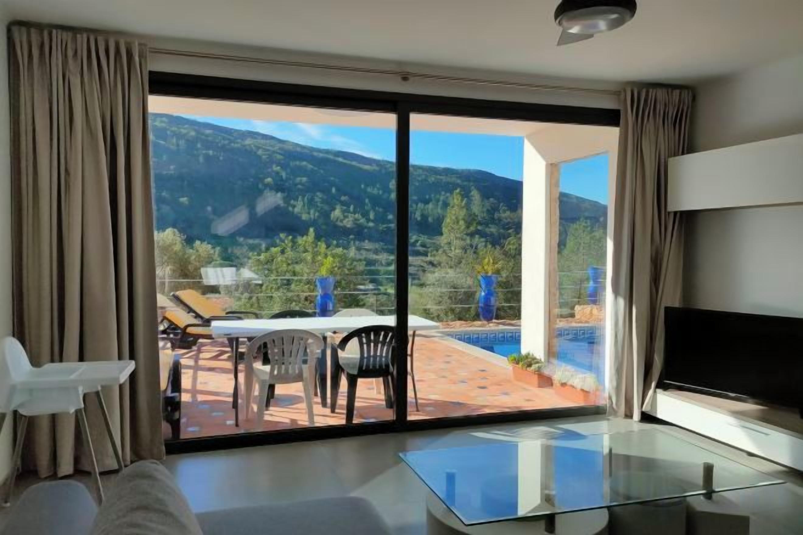 A modern room with a glass table, a sofa, and a TV. It has a sliding glass door leading to an outdoor area with a pool and mountains in the background.