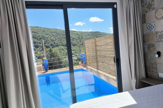 A room with a sliding glass door leading to a pool, a bed, and a view of a hilly landscape.