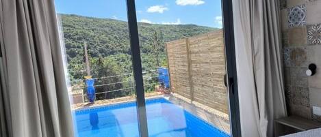 A room with a sliding glass door leading to a pool, a bed, and a view of a hilly landscape.