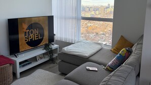 Un salon moderne doté d’une grande fenêtre, d’un téléviseur à écran plat, d’un canapé gris et d’une petite table.