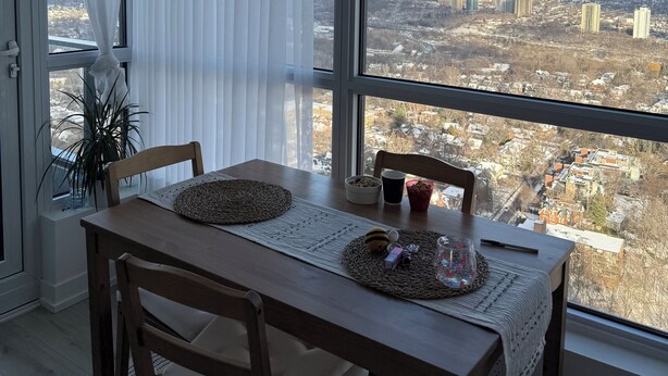 Un coin repas avec une table dressée pour deux personnes, une vue sur le paysage urbain à travers de grandes fenêtres et une plante en pot sur le rebord de la fenêtre.