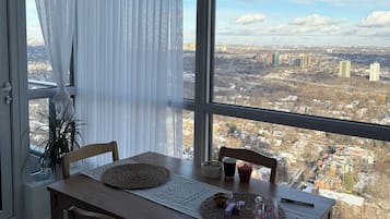 Un coin repas avec une table dressée pour deux personnes, une vue sur le paysage urbain à travers de grandes fenêtres et une plante en pot sur le rebord de la fenêtre.