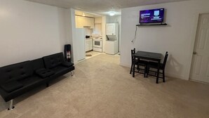 Ein Wohnzimmer mit schwarzem Sofa, einem Esstisch mit Stühlen, einem an der Wand befestigten Fernseher und einer Küche im Hintergrund.