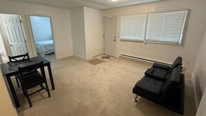 Ein Zimmer mit Esstisch, Stühlen, einem schwarzen Sofa, einer Tür und einem Fenster mit Jalousien.