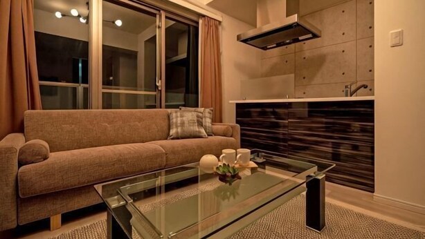Una sala de estar con un sofá, una mesa de centro de vidrio y una zona de cocina con armarios y estufa.