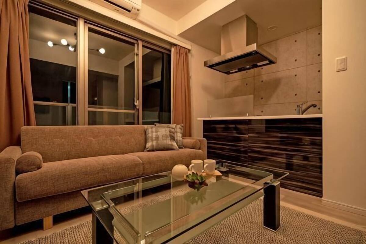 A living room with a sofa, glass coffee table, and a kitchen area with cabinets and a stove.
