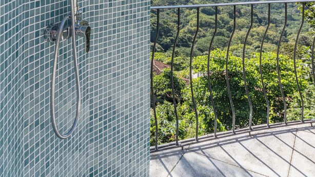 A shower with a rain showerhead and handheld showerhead, a tiled wall, and a balcony with a metal railing overlooking a mountainous landscape. -  Casa Nubes (Puerto Carrillo)