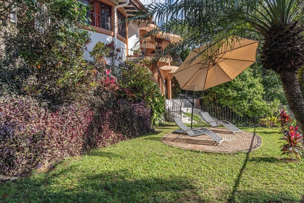 A well-maintained garden with a palm tree, a lounge area with a large umbrella, and a building with balconies in the background.