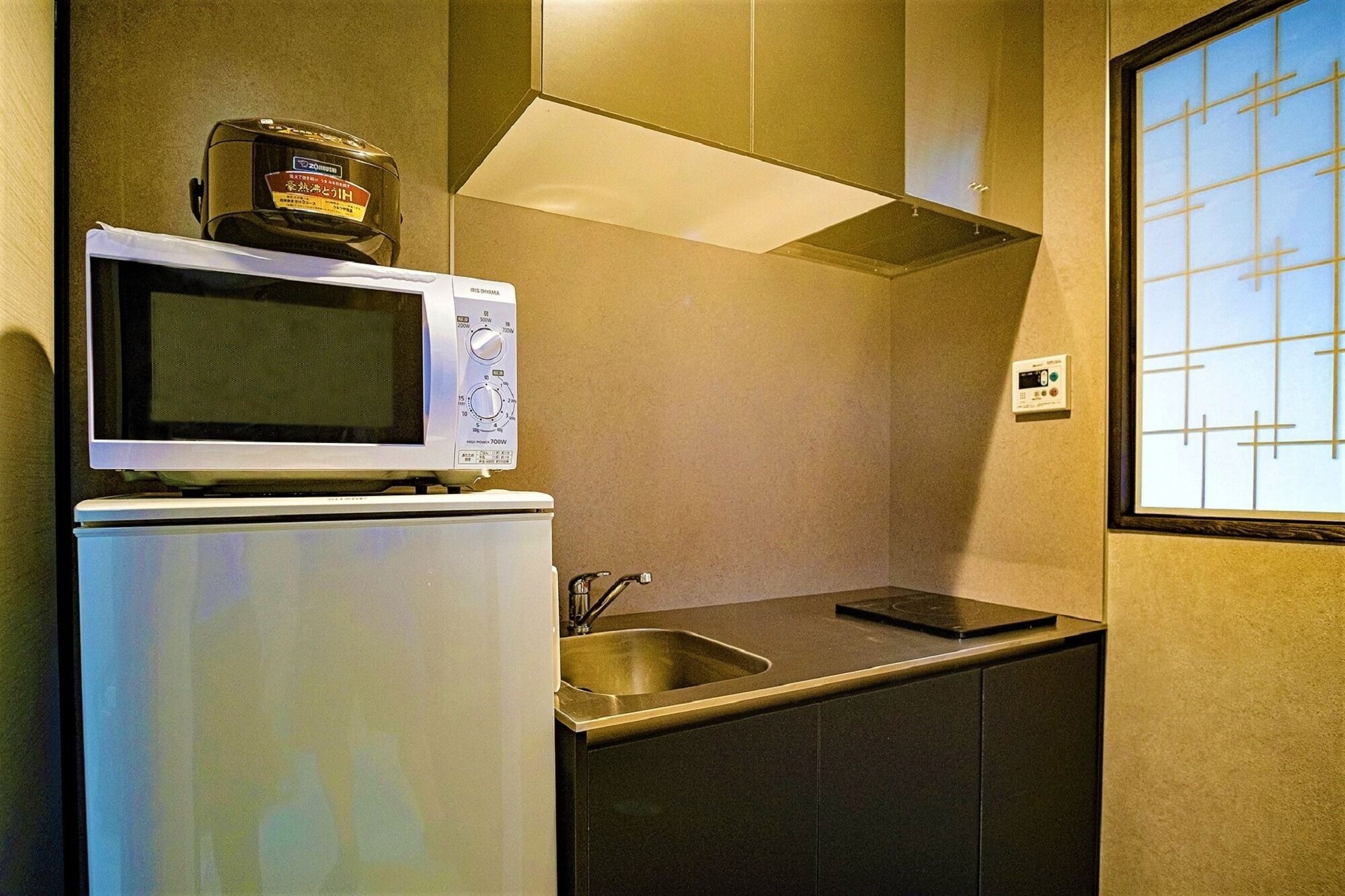 A compact kitchen with a microwave and a small refrigerator, a stainless steel sink, and a window with a grid pattern.