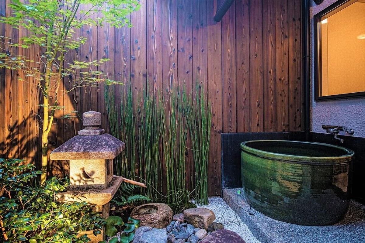Una cerca de madera, un farol de piedra, hierba alta, una maceta verde y un pequeño jardín de rocas.