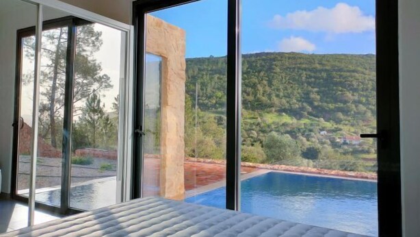 Un dormitorio con un amplio ventanal corredera que da a una piscina y vistas de una ladera boscosa.