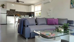 Una sala de estar moderna con un sofá gris, una mesa de centro de vidrio y una cocina al fondo.