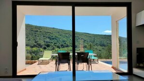 Habitación moderna con puerta corredera de vidrio que da a un patio exterior con mesa y sillas, con vistas a una colina verde exuberante.