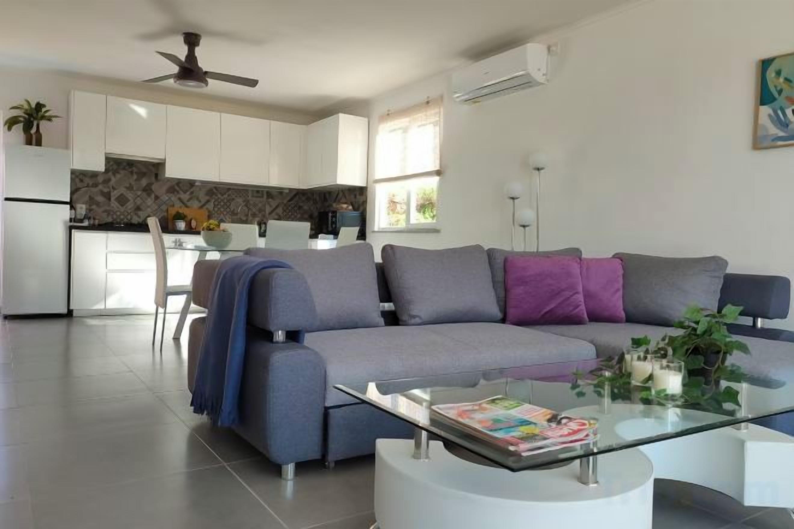 A modern living room with a grey sofa, a glass coffee table, and a kitchen in the background.
