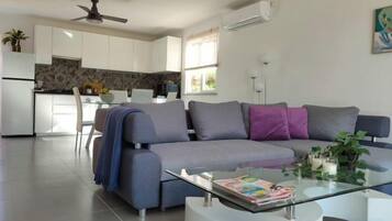A modern living room with a grey sofa, a glass coffee table, and a kitchen in the background.