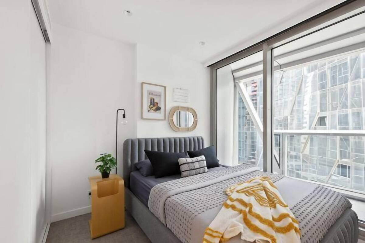 A bedroom with a bed, a bedside table, a window, and a wall-mounted mirror.