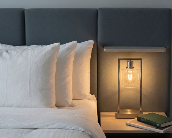 Un lit avec du linge de lit blanc et des oreillers, une table de chevet avec un livre et une lampe.