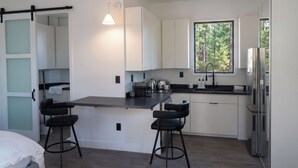 A modern kitchen with a central island, bar stools, a window with a view of greenery, and a refrigerator. - Horizons Sandhills (Patrick)