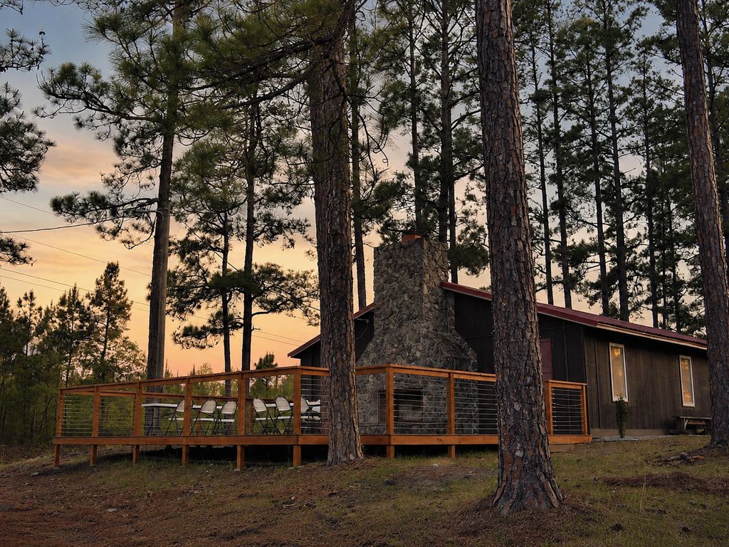 Una cabaña con chimenea de piedra, rodeada de altos pinos, y una terraza de madera con sillas.