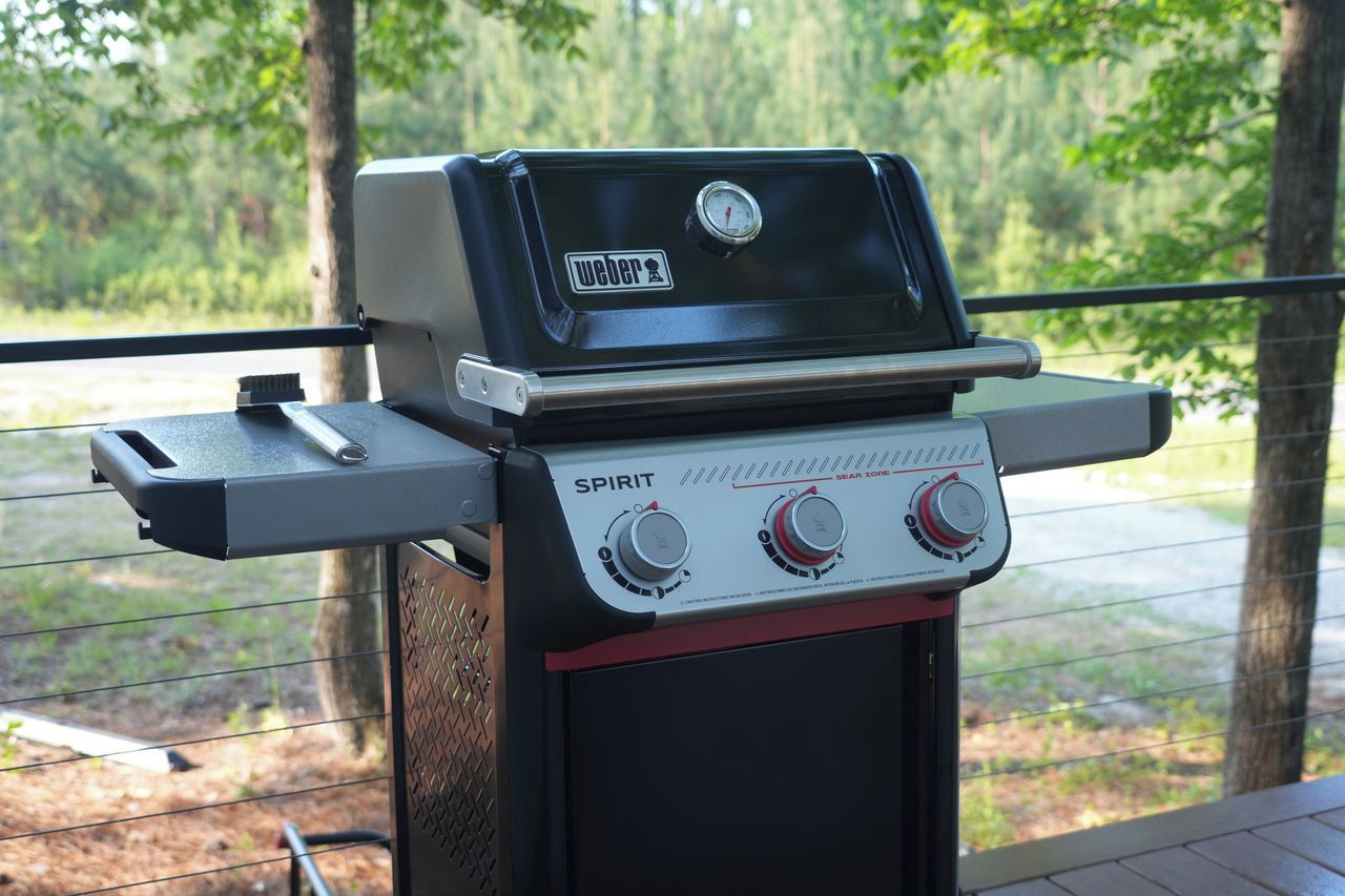 Una parrilla Weber Spirit con pantalla digital y dos perillas de control, con un paisaje boscoso de fondo.