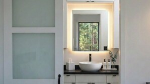 A modern bathroom with a sliding door, a large mirror, a white sink, and a window with a view of trees. - Horizons Sandhills (Patrick)