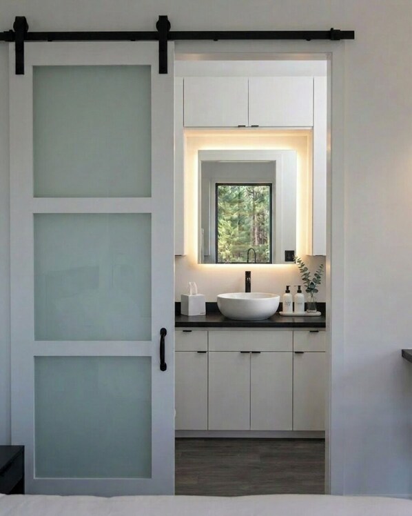 A modern bathroom with a sliding door, a large mirror, a white sink, and a window with a view of trees.