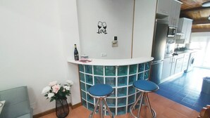 Et køkken med en hvid bordplade, to blå barstole, en dekorativ væg med et vinglas og et hjerte, samt en vase med blomster.