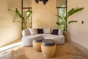 Un salon moderne avec un canapé gris, trois coussins et deux petites tables d’appoint en osier. De grandes plantes vertes sont disposées de part et d’autre du canapé.